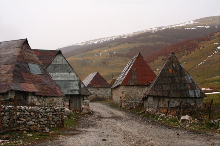 Stone buildings with steep roofs beside a lane in Lukomir, with the mountain hillside behind.